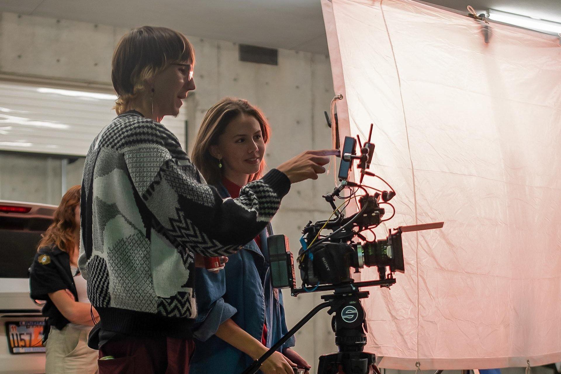 Two Phase 2 Fellows stand in front of a camera, pointing at the screen, with lighting gear behind them.