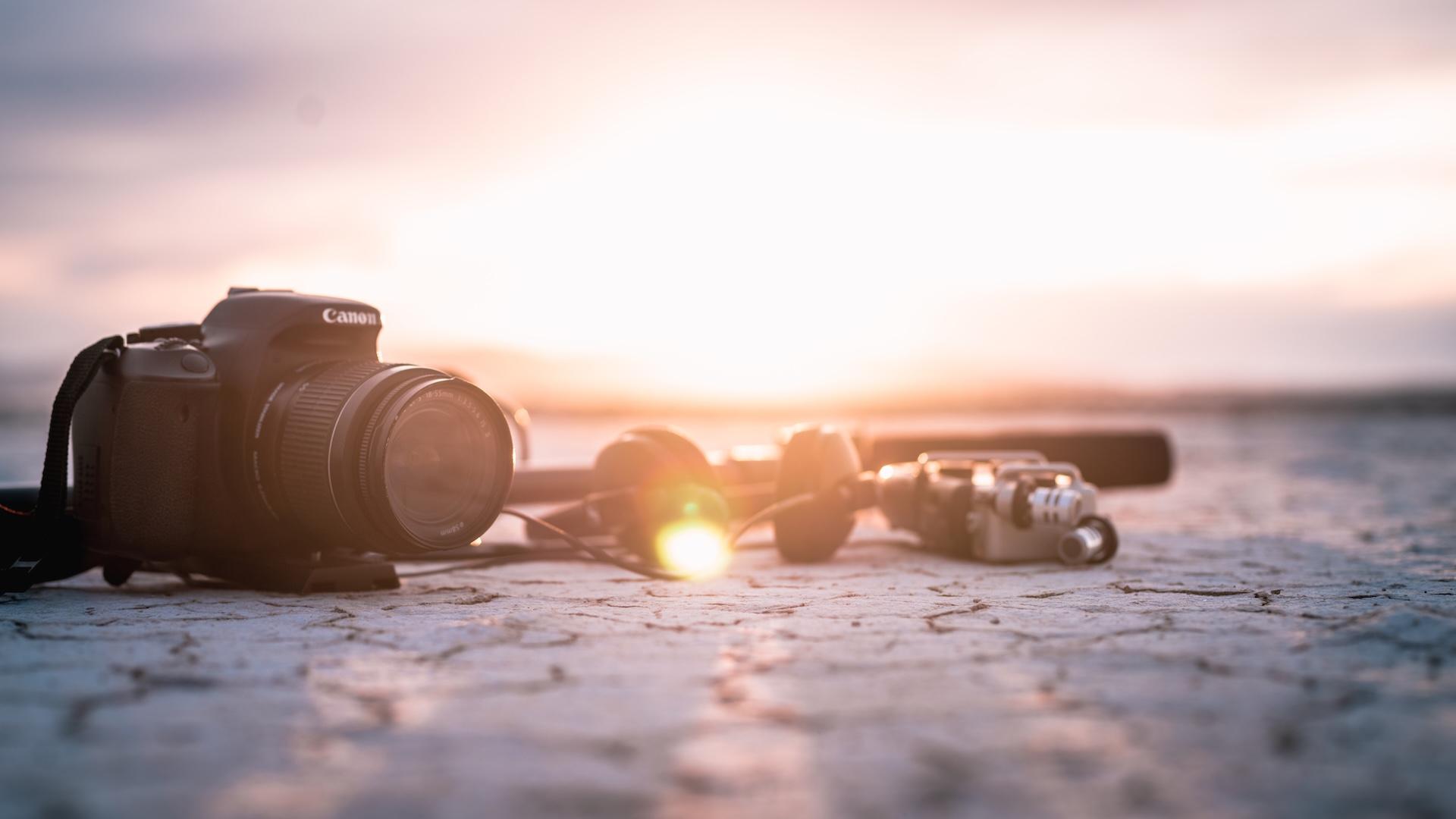 A camera and other film gear sit on a cracked desert ground, the sun setting behind them.