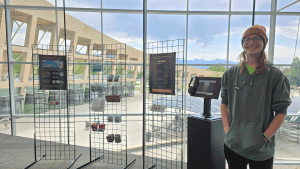 A student stands and smiles in front of an art display, featuring art and zines at the Salt Lake City Public Library.