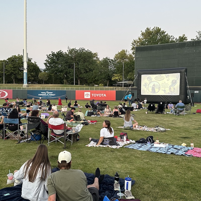 A crowd sits on blankets in a ballpark, watching a film on an inflatable screen.